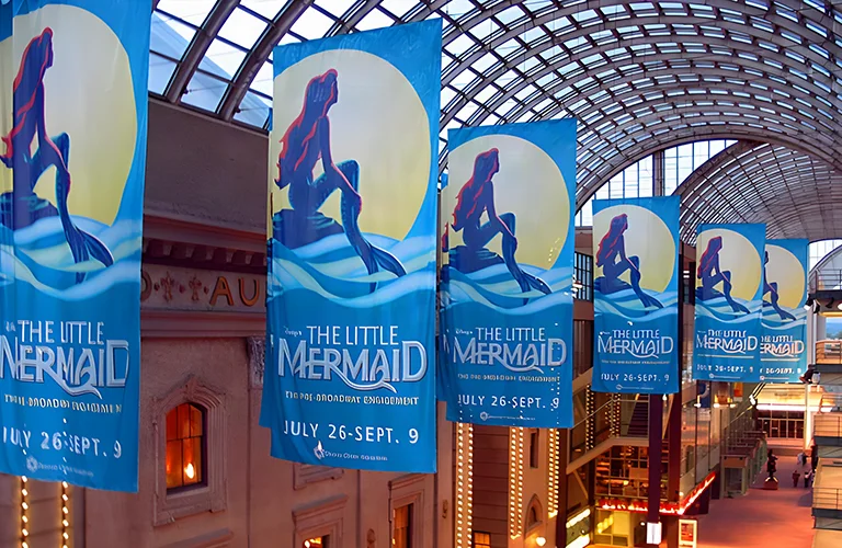 Series of large hanging flags promoting The Little Mermaid displayed inside a glass-roofed indoor venue.