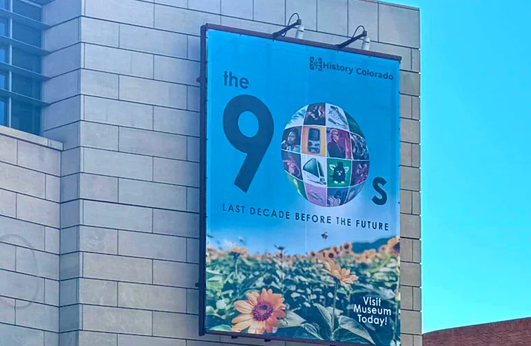 Large outdoor promotional banner for History Colorado’s “The 90s: Last Decade Before the Future” exhibit mounted on the side of a building.