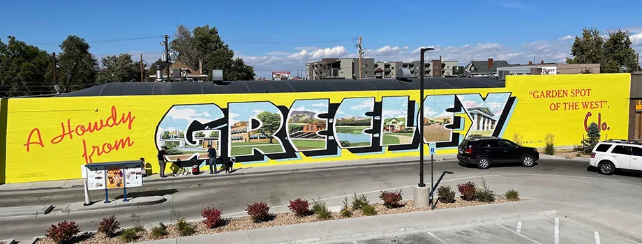 Large outdoor wall mural in Greeley, Colorado featuring colorful postcard-style lettering, local imagery, and the phrase “A Howdy from Greeley.”